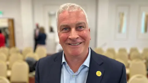 BBC Deputy Andrew Niles - A man with white hair smiling at the camera. He has a blue suit on, with a blue shirt. 