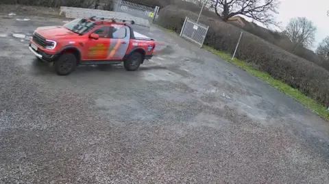 Whetstone Athletic CCTV image of a orange pick up style vehicle driving across a gravel area, with a gate behind