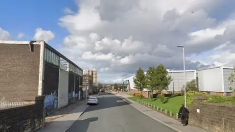A suburban road with a brutalist building and parked car on one side of the road and a grey modern-looking building on the other. A pedestrian dressed in black walks along the pavement on the right.