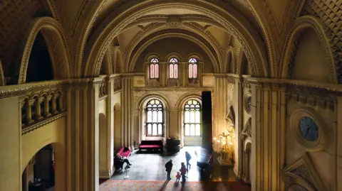 National Trust/John Millar The Gothic grand hall at Penrhyn. The double height room has elaborate arched ceilings, large stained glass windows and large circular clocks carved into the stone 