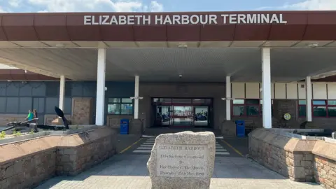 BBC The front of the Elizabeth Harbour Terminal building with a large poritco bearing the name of the building and a stone commemorating the opening of the harbour by Queen Elizabeth II in 1989