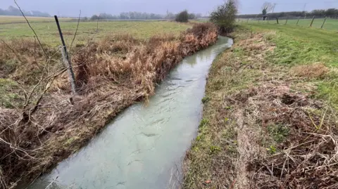 A fast flowing stream is running through an open field there is a water level sensor on the left bank.