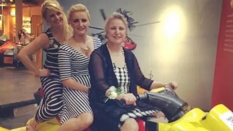 Three woman dressed in black and white dresses - seemingly for an event - all sat together in a row on something that looks like an indoor jet ski ride