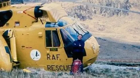Dave Malkinson The picture is blurred because of its age. In the foreground there's a man, Dave Malkinson, wearing a dark blue jumper, leaning against the front of a yellow helicopter. On the side of the helicopter it says RAF Rescue. Malkinson has a red backpack leaning against his legs and has his arms folder. In the background there is a mountain.