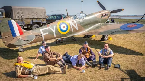 Andy Bell Images A group of people in vintage wartime-style clothing relax on the grass in front of a replica World War Two fighter aircraft. Military vehicles are parked behind the aircraft and the day is sunny