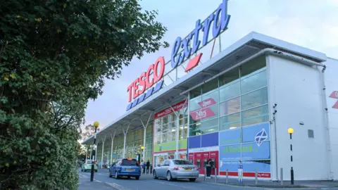 Billy McCrorie A side-on view of a Tesco store with Tesco Extra above the entrance and a few cars outside