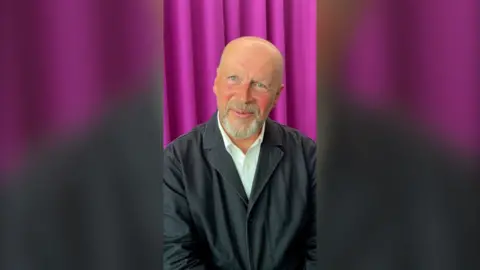A man, James Reed, in front of a purple curtain. He is bald and wears a blazer.