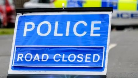 A police road-closed sign with a police vehicle behind it, slightly blurred. The road sign is blue with white writing.