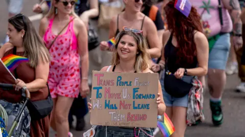 PA Media A Pride parade participant holds a handmade cardboard sign reading, “Trans People Fought To Move Us Forward. We Will Not Leave Them Behind. No LGB Without T.” Other marchers carrying rainbow flags walk behind them.
