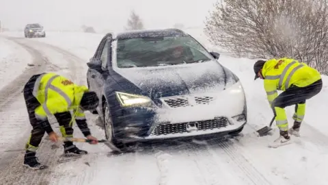 Two men in high vis clothing use spades to dig a blue car out of the now