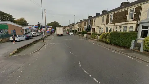 Google Library image of Whalley New Road, Blackburn, showing a shop on the left and terraced houses on the right with parked cars and a van travelling on the road.