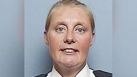 West Yorkshire Police Sharon Beshenivsky smiling at the camera with her blonde hair tied back in a ponytail. She is wearing her police uniform and is stood in front of a grey background. 