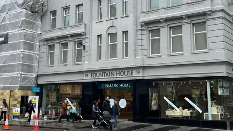 Outside view of Fountain House. It is a large grey building with the Primark Home storefront at the bottom floor. People are walking along the street.