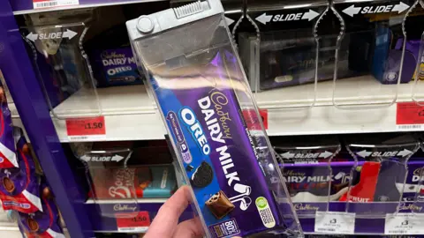 BBC Against a supermarket shelf, we can see a plastic lockbox with a family size bar of Cadbury Dairy Milk Oreo inside. It is a theft prevention measure. The shelves behind show plastic screens on the shelf edge to stop the theft of chocolate in this store. 