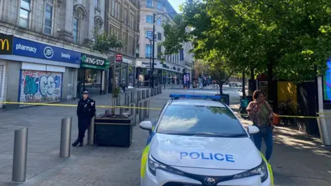 Police car parked in Piccadilly Gardens in Manchester city centre. There is a row of shops and restaurants on the left hand side. A female police officer stands near the police car.