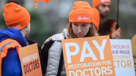 Getty Images A woman with blonde hair and an orange hat with a large orange sign which reads pay restoration for doctors, alongside other people with similar placards.