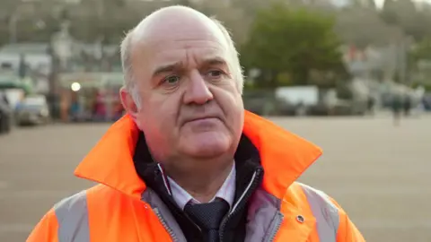 David Rodda wearing a bright orange high-visibility jacket with reflective stripes, standing outdoors in an open paved area. Trees and buildings are visible in the blurred background, along with a few people walking in the distance.