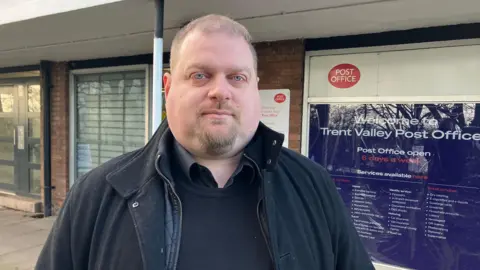 MP Dave Robertson is standing outdoors on a street next to a Post Office. He is wearing a black jumper and black coat over it. He is lookign at the camera.