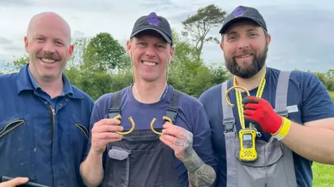 Alan Daniels, Andy Crammond and Richard Forbes are standing together and smiling. The man on the left is bald and has a moustache. The man in the centre is clean shaven and is wearing a black cap, and holding two golden torcs. The man on the right has a beard and is wearing a black cap and holding one golden torc. There are trees in the background.