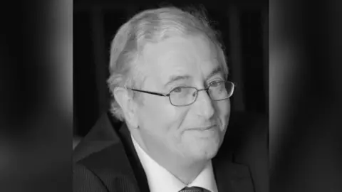 Alzheimer’s Research UK A man with wavy grey hair looks wistfully to camera in a black and white image. He is wearing thin rimmed metal glasses, a dark suit, a white shirt and a grey tie.