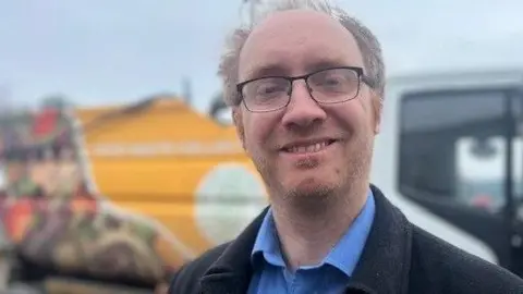 Hull City Councillor Charles Quinn smiling at the camera. This is a head and shoulders photograph and Cllr Quinn is wearing a black coat and a blue shirt. He has short blond hair and is wearing glasses. Behind him is a food waste collection truck.