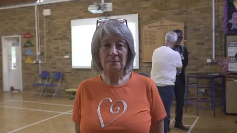 LDRS A woman hair with grey hair and wearing an orange t-shirt with a silver heart motif. She is standing in a school hall and looking at the camera. Two men stand behind her in the background talking to each other.