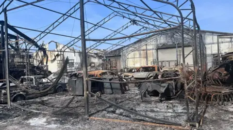 A burnt down warehouse with around six destroyed cars covered in ash, and debris lying around