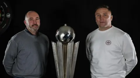 Nodor Lee standing alongside Gerwyn Price with the PDC World Championship trophy in the middle of them. The silver trophy comes up to shoulder height of the men, with a globe on top of it.