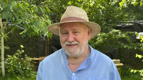 Nimret Kaur / BBC A man, Trevor Williams, wearing a blue shirt and a weaved hat. He is stood in front of some trees.