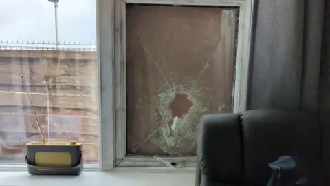 Handout A broken window pane is boarded up with wood. A radio is on a window sill and a chair can be seen nearby.