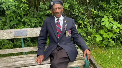 A man wearing a blazer and commemorative medals sat on a park bench surrounded by greenery