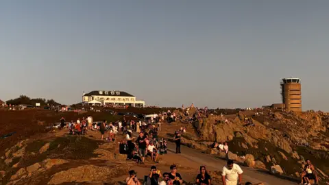 Crowds gather in Corbiere. There is people sat down looking at the sun with glasses