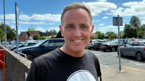 Zac Sherratt/BBC A man in a black tops stands in a car park, smiling into the camera.