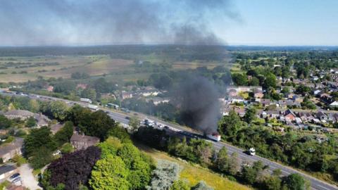 Aerial image of a lorry stopped at the side of an A-road, with flames visible on top and a plume of black smoke billowing into the air