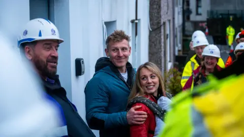 South Shore A group of people, many in hard hats and hi-vis jackets, stand outside a row of houses. In the centre of the image, a smiling man in a blue jacket has his arm around a blonde woman in a red jacket wearing a baby-carrier strapped to her front. DIY SOS presenter Nick Knowles, a bearded man wearing a white hard hat,  stands on the far left of the image.