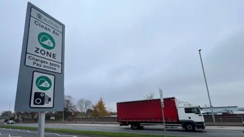 BBC/Jamie Coulson A road sign indicates Bradford's Clean Air Zone. It is captioned "Charges apply, pay online". A red lorry drives along the dual carriageway in the background.