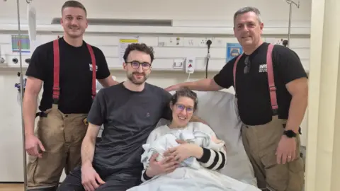Two firefighters in a black T-shirt and khaki trousers stood on either side of the bed. A couple hugging a baby are sat on a bed smiling. The dad is wearing a grey T-shirt and dark trousers while the mum is wearing round glasses and a white jumper.