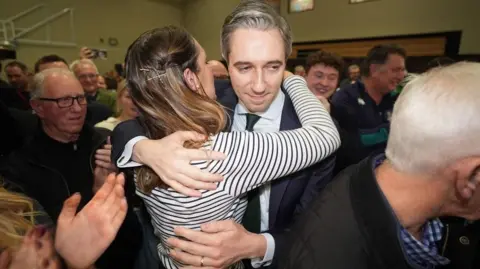 PA Media Simon Harris who has greying hair parted to the side and is wearing a dark side is hugged by his wife who has long brown hair and is wearing a striped top at an election count centre