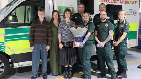 Chloe Oakley-Mudge is pictured with her family and ambulance staff standing in front of an ambulance. Her brother and mother are in the photo as well as two female and three male ambulance staff. One of the female staff is holding a large bunch of flowers in a box
