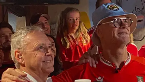 Dieter Steinbusch Mike and Dieter surrounded by Wrexham fans looking up most likely at a television as a Wrexham game is on. They are wearing Wrexham tops. And Dieter has a blue hat on.