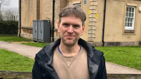 BBC Henry Winchester sat on a bench. He has short dark hair and stubble. He is wearing a black coat and a brown jumper. He is sitting in front of a brown stone building with a path and grass.