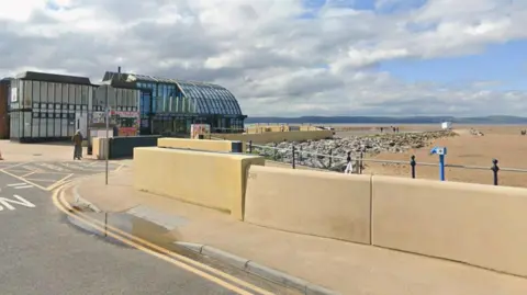 A view of the boathouse building at West Kirby beach with a yellow beach wall, sandy beach and a view of Welsh hills in the distance.