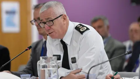 Jon Boutcher, he has white hair and glasses and is wearing a white shirt shirt with PSNI emblems. He is sitting at a table and is leaning forward speaking into a black microphone. A jug of water is in front of him. 