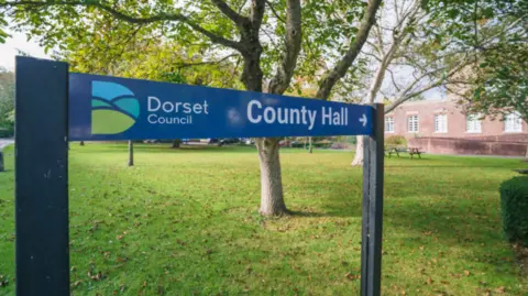 A Dorset Council sign pointing towards County Hall in Dorchester, with County Hall in the distance.