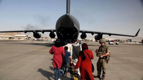 Woman and children with their backs to the camera walk past a soldier as they board a RAF aircraft at Kabul Airport in 2021