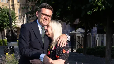 PA Media A man in a dark suit, wearing glasses and smiling broadly hugs a blond woman in a black floral dress on a sunny day outside the houses of parliament. 