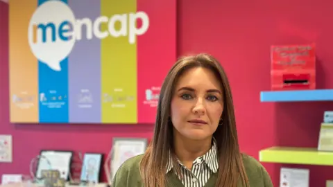 Karen Gilgunn in stood in an office. She had long brown hair and is wearing a green and white striped shirt, with a matching green jumper over the top.
The walls are bright pink and shelves are blue and yellow.
The Mencap sign is striped and multi-coloured.