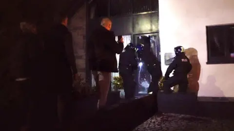 Three officers wearing helmets and protective suits gathered round a glass door of a house while three plain clothed officers stand in the shadows behind them further down a path.