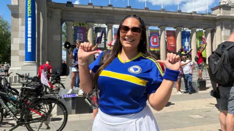 Layla is pictured wearing a Bosnian jersey and a white skirt with sunglasses. She has long black straight hair. Behind her are the gates to Toronto's stadium grounds, decorated with Fifa 2026 banners.