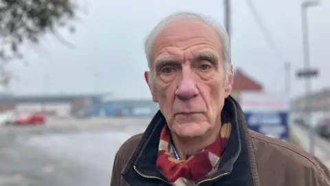 Elizabeth Baines/BBC A man with white hair stands in front of Featherstone's Rugby ground. He wears a brown coat with a burgundy and mustard scarf